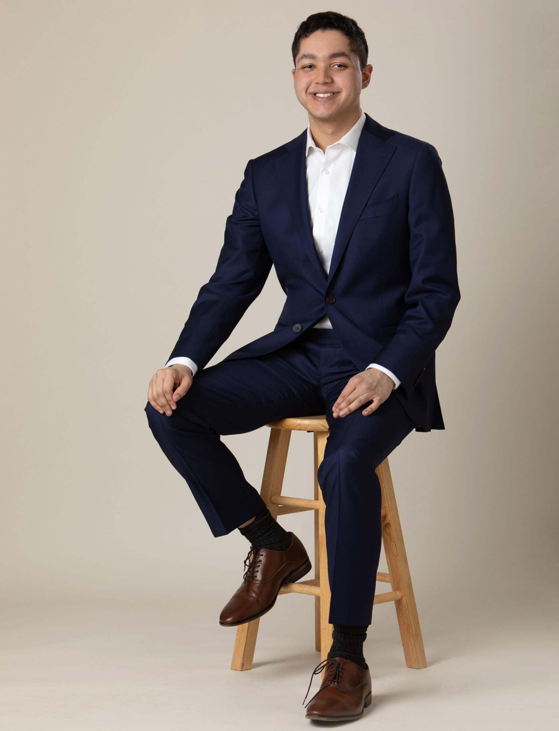 Picture of Kevin Johnson-Cadle smiling in a navy blue suit with a white shirt; while sitting on light brown stool.